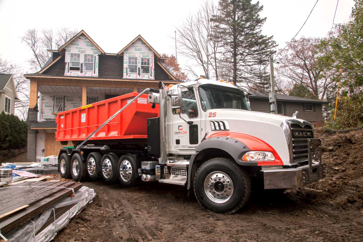 view of a Dem-Con truck dropping off a dumpster at a new house build site